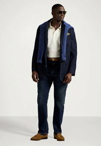 Bărbat purtând ochelari de soare, sacou bleumarin cu pulover albastru aruncat pe umeri, cămașă polo albă, blugi închiși la culoare, mocasini maro și curea maro.