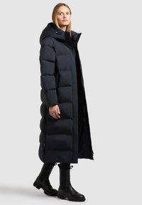 Langer navyblauer Puffer-Mantel mit Kapuze, ausgestattet mit einem Reißverschluss, großen gesteppten Abschnitten und zwei seitlichen Taschen. Dazu schwarze Stiefel.