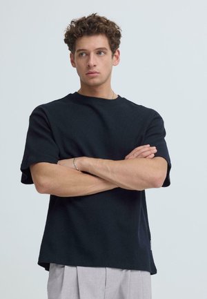 Jonge man met krullend haar, gekleed in een zwart shirt met korte mouwen en lichtgrijze broek, staat met gekruiste armen en kijkt naar de zijkant.