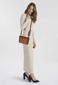 Braune Leder-Crossbody-Tasche mit einem langen Riemen, rechteckiger Form und einem dekorativen runden Metallbesatz. Getragen mit einem langen, cremefarbenen Kleid.