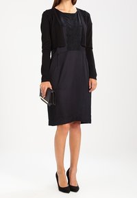 Svart klänning med en strukturerad bodice, knälång, lång ärm, i kombination med en svart cardigan och högklackade skor. Hållande en liten svart kuvertväska.