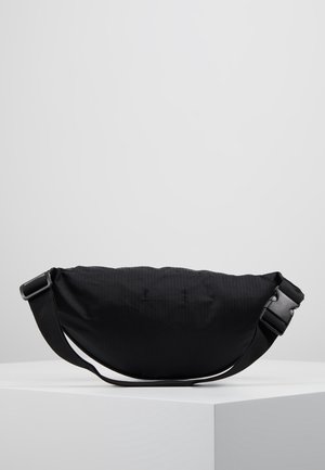 Czarna materiałowa torba na biodra z regulowanym paskiem i plastikową klamrą, prezentowana na białej platformie na tle jednolitego jasnoszarego tła.