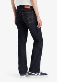 Personne portant un jean droit bleu foncé avec des coutures orange et des baskets bleues avec des accents blancs, debout devant un fond blanc.