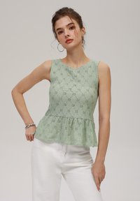 Zaļš mežģīņu peplum tops ar ziedu izgriezumiem, noapaļota kakla izgriezuma forma un bez piedurknēm. Apvienots ar baltām biksēm.