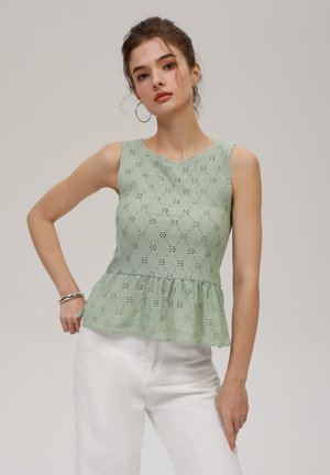 Green lace peplum top with floral cutout pattern, rounded neckline, and sleeveless design. Paired with white pants.