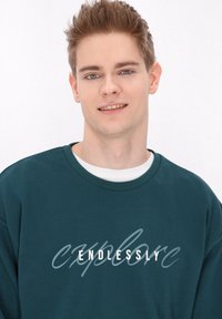 Tmavě modrý svetr s kulatým výstřihem, s výrazem "ENDLESSLY" v tučném bílém písmu a kurzívou. Měkká tkanina a uvolněný střih.