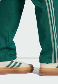 Gröna träningbyxor med tre ljusgröna ränder; beige skor med gröna ränder, texturerad ovandel och gummigummi-sulor, med varumärke.