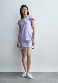 Νεαρή γυναίκα με ανοιχτό μωβ φόρεμα με κοντά μανίκια και λευκά αθλητικά παπούτσια στέκεται μπροστά σε ένα απλό ανοιχτό γκρι τοίχο, κοιτάζοντας προς το πλάι.