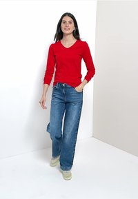 Röd långärmad v-ringad topp kombinerad med vida blå jeans och ljusa skor. Enkel design, enfärgat, avslappnad passform.