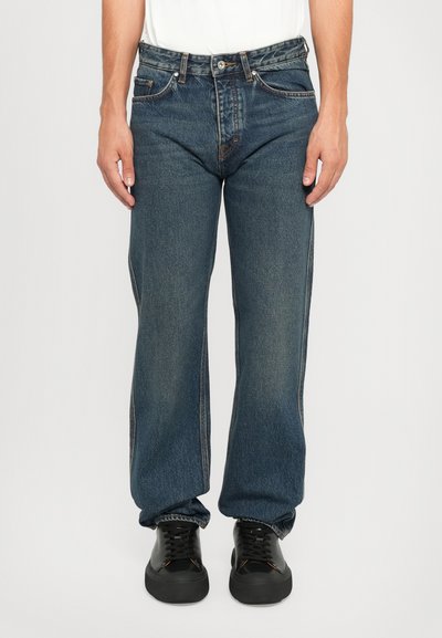 Dark blue straight-leg denim jeans with five-pocket design, contrasting stitching, and a worn look, paired with black sneakers.