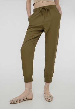 Persona che indossa pantaloni verde oliva con coulisse e polsini elasticizzati e sandali beige piatti con cinturini, in piedi con le mani nelle tasche.
