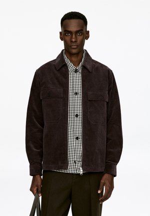 Dunkelbraune Cordjacke mit zwei Brusttaschen und einem Reißverschluss vorne. Darunter ein schwarz-weißes Karoshemd mit Knopfleiste.