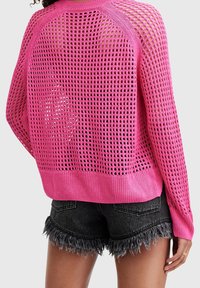 Różowy sweter dziany z luźnym, siatkowym wzorem, szerokim dekoltem i ściągaczami na rękawach, zestawiony z postrzępionymi czarnymi szortami dżinsowymi.