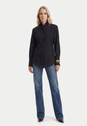 Frau steht und trägt ein eng anliegendes schwarzes langärmeliges Oberteil, blaue Jeans, braune Stiefel und ein großes goldenes Armband am linken Handgelenk.