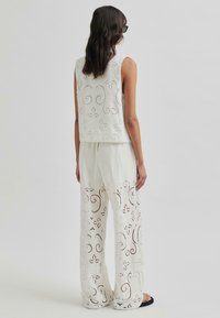 Weißes Spitzenoberteil und weit geschnittene Hose mit aufwändigen, ausgeschnittenen Blumenmustern. Weicher Stoff mit lockerem Schnitt und gewelltem Saum. Schwarze Sandalen.