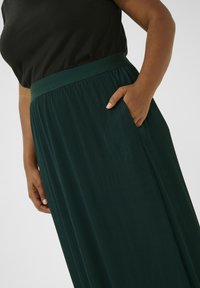 Falda verde plisada con una amplia cinturilla elástica y bolsillos laterales, hecha de una tela suave, con rayas verticales para dar textura.