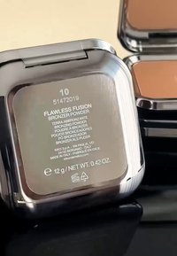 Kompaktowy puder brązujący w metalicznej srebrnej obudowie z przezroczystą pokrywką, z informacjami o produkcie i numerem odcienia umieszczonym z tyłu.