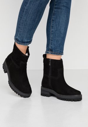 Bottines noires en daim avec fermeture éclair latérale et semelles crantées, portées avec un jean skinny bleu retroussé sur un sol clair.