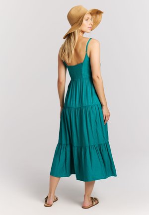 Vrouw die een teal mouwloze midi-jurk draagt, een strooien hoed met brede rand en platte sandalen, staand en over haar schouder kijkend.