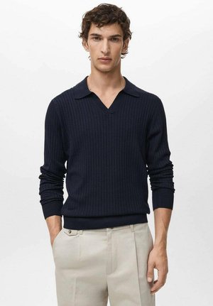 Young man wearing a navy ribbed long-sleeve sweater with a collar and beige trousers, standing with one hand in pocket against plain background.