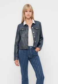 Skjortekort denimjakke i mørkeblå med sølvmuttere, to brystlommer og kontrastsøm, kombinert med en hvit bluse og stripete bukser.