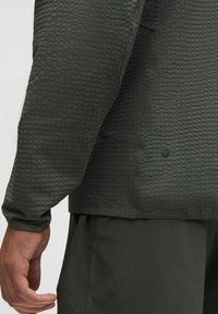 Mørkegrøn, tekstureret langærmet bluse med et geometrisk mønster og et rundt logo i kanten, parret med matchende shorts.