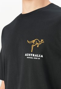 Svart t-skjorte med gul kenguru-grafikk og hvit tekst "AUSTRALIA RIP CURL SURF CO" på brystet.