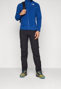 Man i blå fleecejacka, svarta utomhusbyxor och gröna vandringsskor som håller i en svart ryggsäcksrem mot en enkel bakgrund.