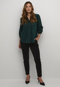 Tirkizna bluzica z razpihanimi rokavi, okrašena z odprtimi detajli in V-izrezom. Kombinirana z črnimi tesnimi hlačami in črnimi špicastimi petami.