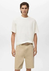 En ung mand står mod en ensfarvet baggrund, iført en hvid kortærmet skjorte og beige shorts med et brunt bælte om taljen.