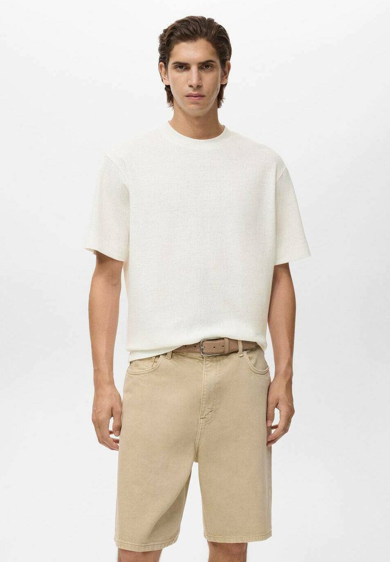 En ung mand står mod en ensfarvet baggrund, iført en hvid kortærmet skjorte og beige shorts med et brunt bælte om taljen.