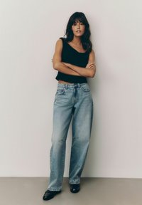 Zwart cropped top en lichtblauwe wijde jeans met een vervaagde afwerking. Outfit compleet met zwarte schoenen. Model poseert tegen een effen achtergrond.