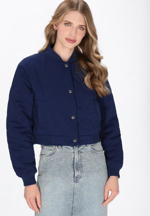 Vrouw met lang golvend blond haar draagt een marineblauwe gewatteerde jas met drukknoopsluitingen en een hoogzitterlichte spijkerrok tegen een witte achtergrond.