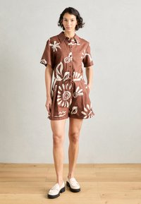 Abito camicia corto marrone con maniche corte, motivo floreale bianco, vita arricciata e lacci. Abbinato a scarpe crema e indossato su un pavimento in legno chiaro.