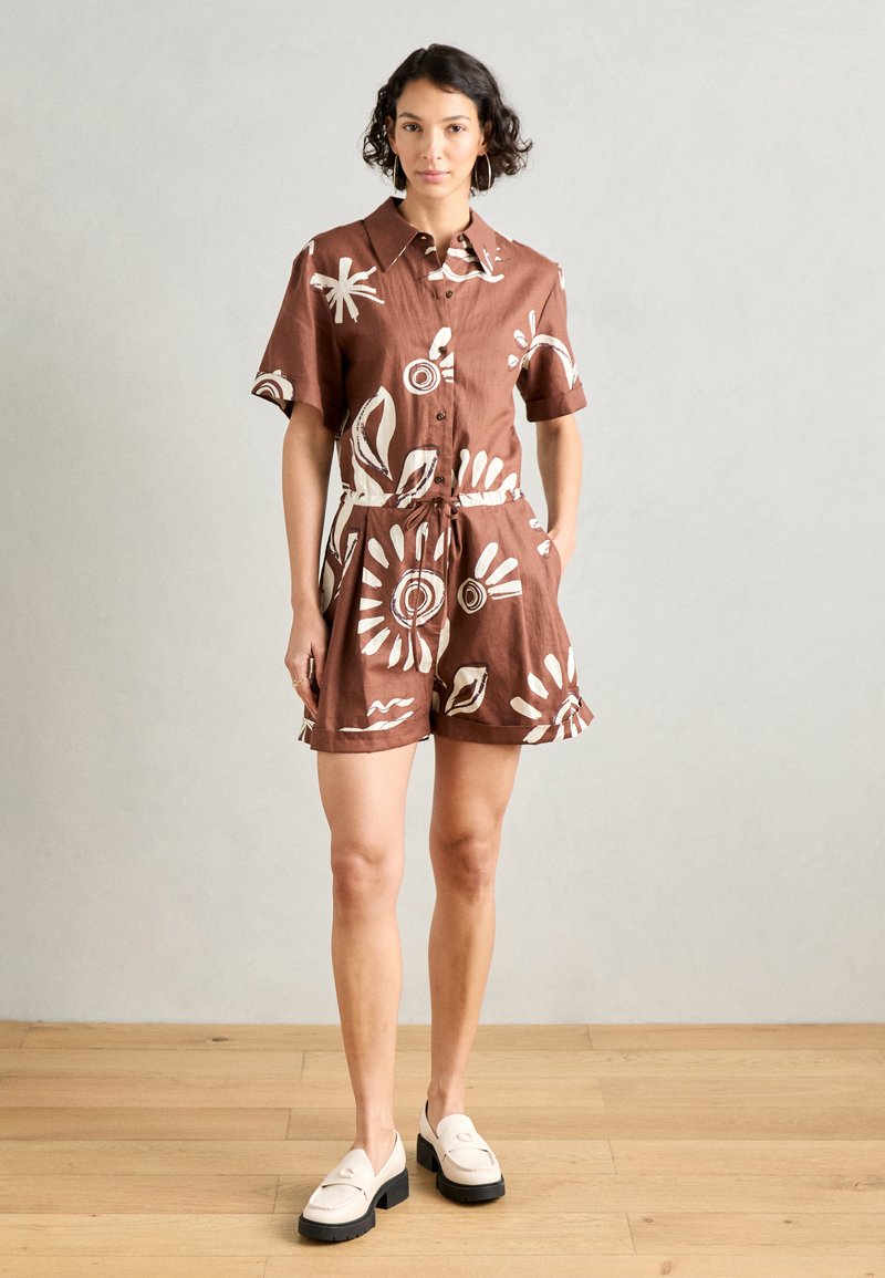 Abito camicia corto marrone con maniche corte, motivo floreale bianco, vita arricciata e lacci. Abbinato a scarpe crema e indossato su un pavimento in legno chiaro.