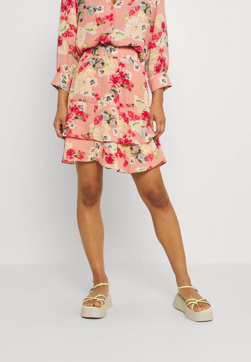 Květinová růžová sukně s vrstveným designem, zdobená červenými, zelenými a modrými vzory; doplněná platformovými sandály s béžovým základem a žlutými popruhy.