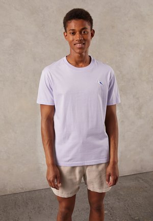 Helle lila Baumwoll-T-Shirt mit Rundhalsausschnitt, mit einem kleinen blauen Vogel-Logo auf der linken Brust. Modell trägt beigefarbene Cordshorts.