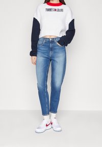 Suéter recortado blanco y marino con diseño de punto de cable; vaqueros de cintura alta azules; zapatillas blancas con acentos rojos; modelo de pie mirando hacia adelante.