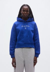Modrá skracovaná mikina s kapucňou so sťahovacou šnúrkou a vyšitým logom značky na hrudi. Kombinovaná so sivými teplákmi. Hladká textúra.