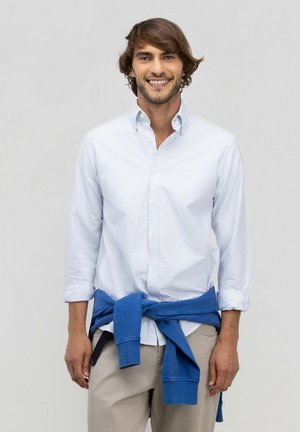 Hombre sonriente con cabello castaño de longitud media que lleva una camisa blanca abotonada y una sudadera azul atada alrededor de su cintura.