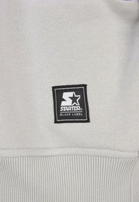 Szara tkanina dresowa o miękkiej fakturze, z czarną kwadratową metką z białym logo "STARTER" i napisem "BLACK LABEL".