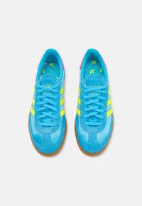 Adidași din suede albastru cu dungi galbene, talpă din cauciuc și accente roz. Au o suprafață texturată și o branț cu un model și logo-uri.