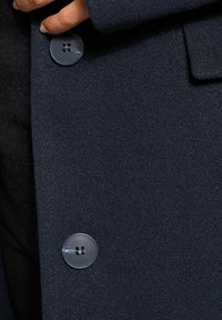 Tessuto di un cappotto blu navy scuro con una finitura texture, dotato di un grande bottone rotondo e cuciture visibili. Mano che afferra il tessuto vicino al bottone.