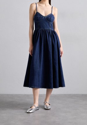 Žena v tmavě modrých denimových midi šatech s tenkými ramínky a stříbrných Mary Jane balerínkách stojí na šedé podlaze před obyčejnou zdí.