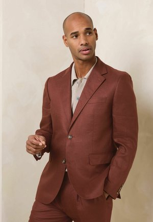 Férfi rozsdabarna öltönyben és bézs, galléros ingben, semleges, texturált háttér előtt áll, kissé oldalra nézve.