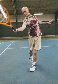 Teniso žaidėjas dėvi trumpomis rankovėmis būdingo smėlio spalvos ir granaatinio rašto polo marškinėlius, smėlio spalvos šortus ir baltus sportinius batus, laikydamas oranžinę raketę ant aikštelės.