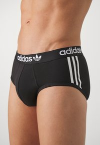 Sorte atletiske briefs med hvid Adidas-logo og tre striber på livet; tætsiddende design; glat, strækbart stof.