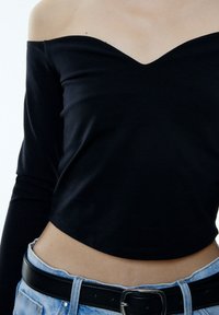Schwarzes, schulterfreies Langarmoberteil mit V-Ausschnitt, kombiniert mit hellblauen, hochgeschnittenen Jeans und einem schwarzen Gürtel mit einer silbernen Schnalle.
