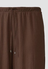 Bruine textuur broek met een elastische tailleband en een trekkoord. De stof heeft een subtiel ruitpatroon en glanzende hardware accenten.