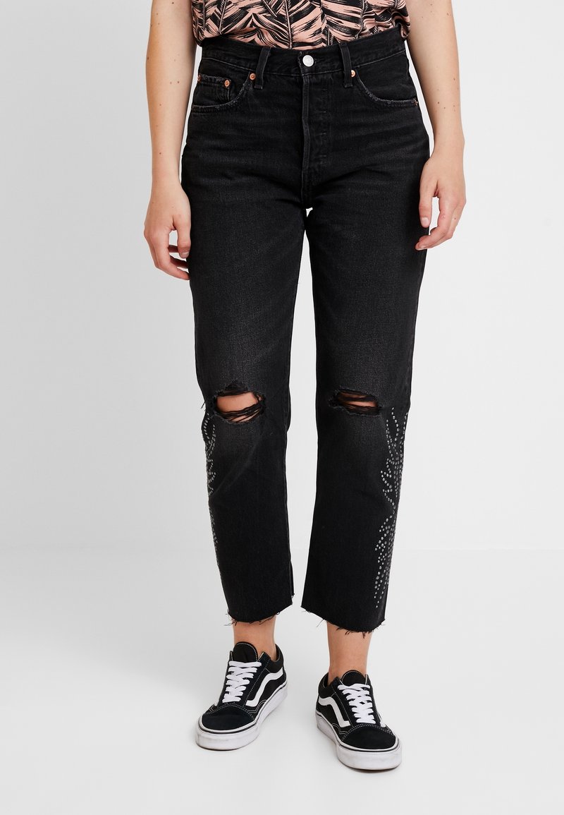 Personne portant un jean noir déchiré avec des motifs en strass sur les jambes et des baskets noir et blanc, posant devant un fond uni.